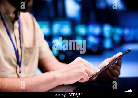 System administrator using tablet to examine server rows for power fluctuations, ensuring uninterrupted computing power. Manager monitoring high tech facility hardware, close up Stock Photo