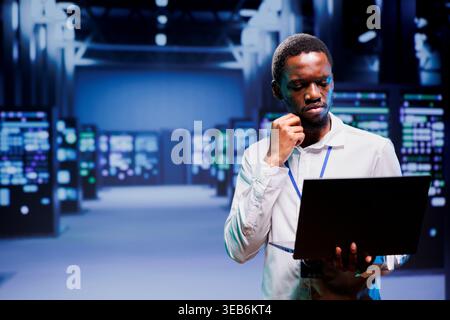 Electrician inspecting blade servers delivering online web content to customers. African american wireman using laptop to take note of critical systems, looking for damaging errors Stock Photo
