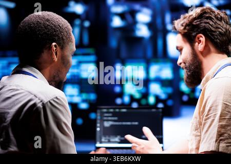 IT developer collaborating with coworker in data center, crafting script code on laptop terminals. Proficient programmers engaging in maintenance work, ensuring optimal functionality and efficiency Stock Photo