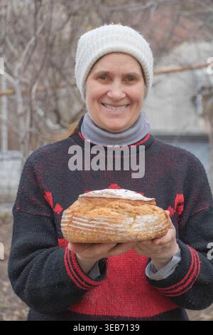 Smiling woman in a red and black patterned sweater and white knit hat holding a fresh loaf of artisanal sourdough bread outdoors Stock Photo