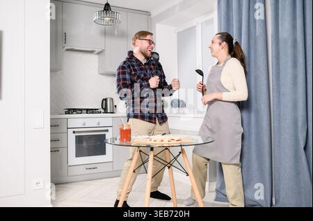 Cheerful young couple singing into kitchen spatulas like microphones, laughing while cooking pizza at home. Happy man, woman having fun, dancing, joki Stock Photo
