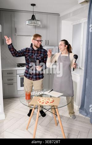 Happy young couple dancing, having fun while cooking pizza together in modern kitchen. Cheerful man, woman in apron playing with kitchen utensil. Dome Stock Photo
