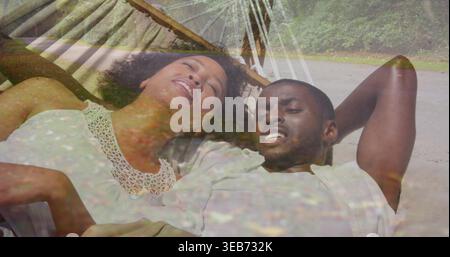 Relaxing couple wearing lace top, open shirt in woven hammock over sand, ropes, posts, leaf overlay Stock Photo