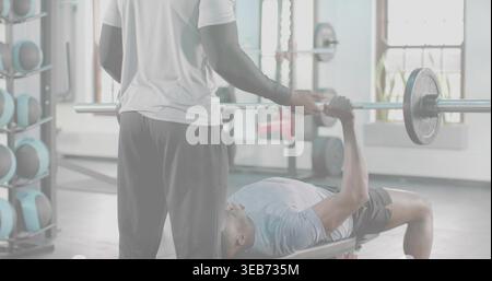 Spotting trainer in white shirt supporting lifter in blue shirt on bench in weight room, barbell. Gym, strength, training, muscular, athletic, coachin Stock Photo