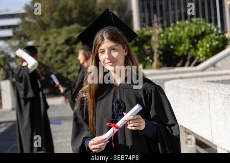 Female graduate standing on campus plaza wearing black gown and mortarboard, holding rolled diploma Stock Photo
