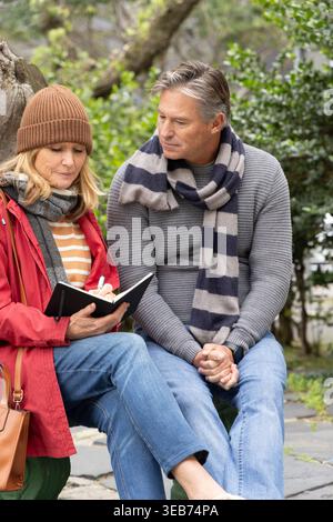Couple sitting on stone ledge in park, woman wearing beanie and scarf writing in black notebook Stock Photo