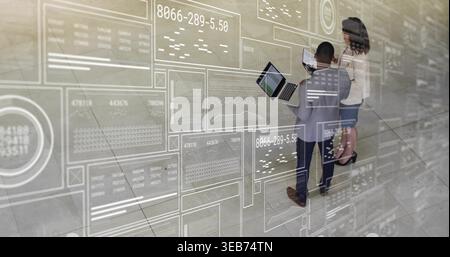 Discussing two professionals in suits using laptop, tablet in atrium with data overlay, copy space Stock Photo