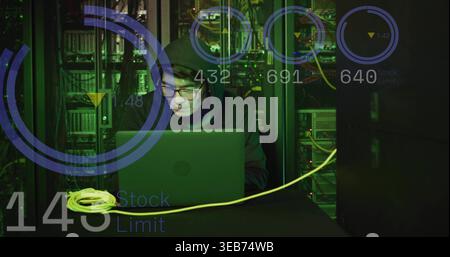 Sitting Korean tech in hoodie monitoring laptop in server room, with yellow Ethernet cable and HUD Stock Photo