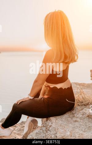 Sunset ocean woman sits on a rocky cliff at the beach watching the horizon during golden hour for meditation and mindfulness Stock Photo