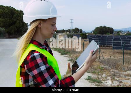 Woman engineer in safety helmet and vest checks performance data on a tablet at a solar farm, monitoring photovoltaic systems for sustainable, clean e Stock Photo
