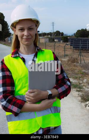 Female engineer or technician with a white hardhat and safety vest holding a tablet. Standing in front of a solar panel farm and electricity pylon. Sy Stock Photo