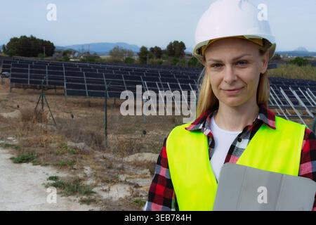 Woman engineer in a hard hat and safety vest holding a tablet, standing in front of rows of solar panels at a photovoltaic power station, symbolizing Stock Photo