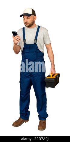 Handyman in blue overalls holding toolbox and using smartphone. Professional repairman checking orders on phone. Stock Photo