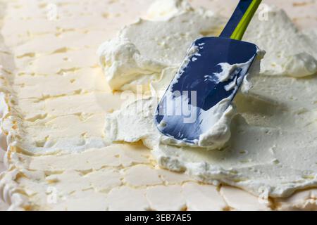 Close-up of a pastry chef's hand using a silicone spatula to spread thick white cream over a baked meringue layer Stock Photo