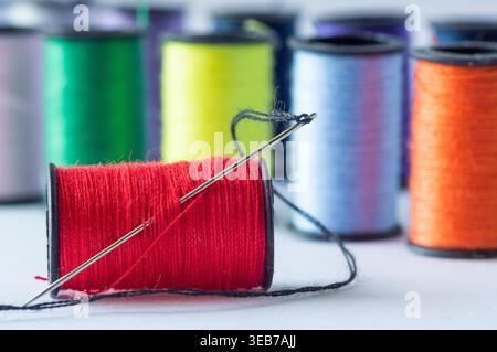 A spool of red thread with a needle, set against a background of spools of colored thread on a white background Stock Photo