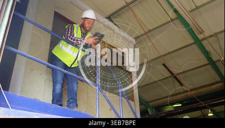Leaning site worker checking tablet on platform at warehouse, white hardhat, neon vest, copy space Stock Photo