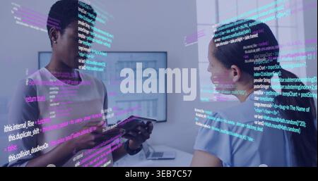 Holding tablet, left woman in workwear reviewing AR code while peer listening in office, monitors Stock Photo