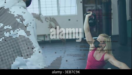 Pressing woman in pink top wearing black face mask raising kettlebell overhead at gym with overlay Stock Photo