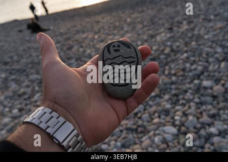 Batumi, georgia 21 april 2026: hand holding painted pebble with smiling face and wave motif on batumi black sea beach — souvenir keepsake from seaside Stock Photo