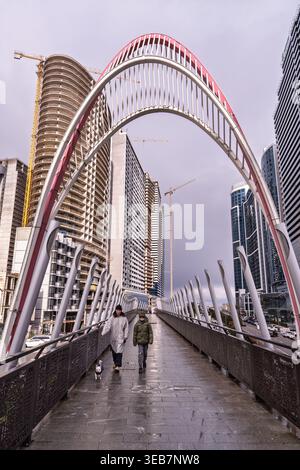 Batumi, georgia 21 april 2026: person and child walking a small dog on a modern pedestrian arch bridge, with contemporary urban skyline and constructi Stock Photo
