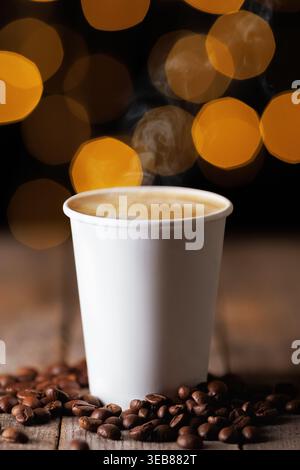 Steaming hot coffee in plain white paper cup on wooden table, with scattered roasted coffee beans and warm bokeh lights Stock Photo