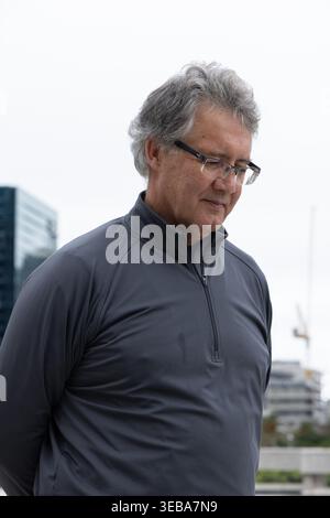 Senior man standing centered on urban plaza, wearing dark zip pullover and eyeglasses, crane behind Stock Photo