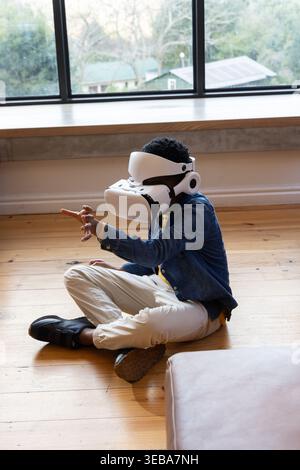 African American boy sitting on wooden floor wearing white VR headset and denim jacket by window Stock Photo