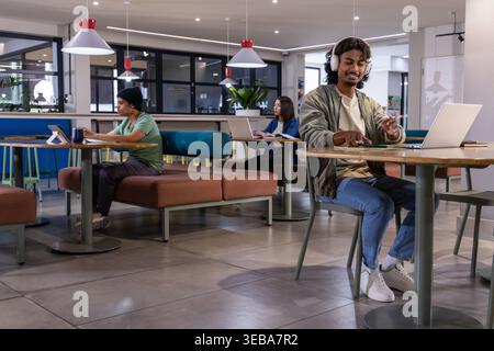 Diverse freelancers with Indian man wearing headphones using laptop in coworking cafe, copy space Stock Photo
