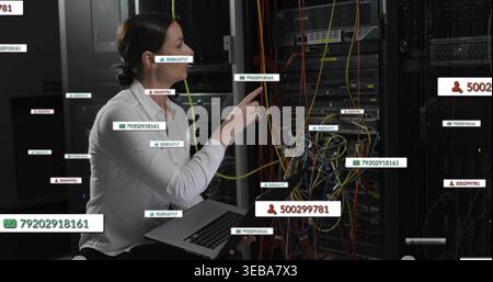 Kneeling IT tech in white blouse touching patch panel with laptop in datacenter, showing cables Stock Photo