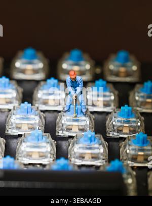 tiny scale model worker in a blue uniform and orange hard hat uses a tool on a blue mechanical keyboard switch, symbolizing tech support, hardware rep Stock Photo
