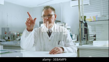 Pointing senior scientist sitting at lab bench, wearing lab coat and glasses, with monitor Stock Photo