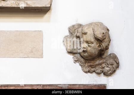 Worn stone cherub relief mounted on a white wall, showing detailed carving, aged texture and architectural ornamentation. Close-up of a historic decor Stock Photo