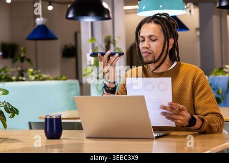 African American man in mustard sweater speaking on phone, holding chart, using laptop, copy space Stock Photo