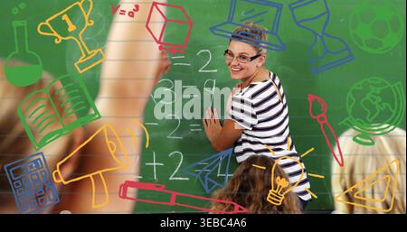 Teaching teacher in glasses striped top pointing at green chalkboard with icons and raised hands Stock Photo