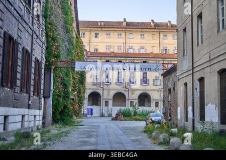 A cobblestone alleyway leads to a historic courtyard in Turin, Italy, with a white banner reading 'LA CAVALLERIZZA È PER TUTTI' spanning between old Stock Photo