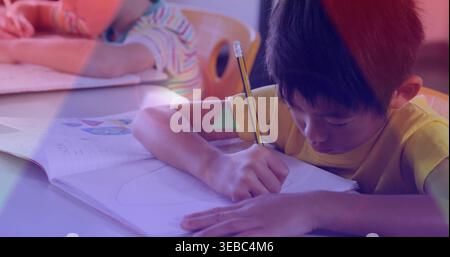 Leaning boy in yellow T-shirt writing in elementary classroom, holding pencil over open workbook Stock Photo