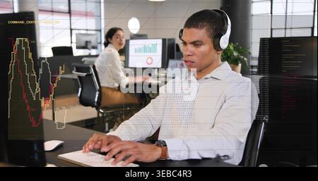 Typing man wearing headset, shirt in open office, using keyboard and mouse, screens showing data Stock Photo
