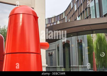 A close-up view of a large, glossy red sculptural object resembling a giant coffee pot or moka pot in the foreground. Behind it stands a modern Stock Photo