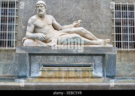 A weathered marble statue of a reclining male figure, representing a river god, positioned on a stone pedestal above a trickling fountain. The Stock Photo