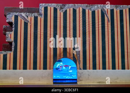 A close-up view of an art installation featuring a wall with peeling striped wallpaper in cream, dark blue, and red vertical lines. The wallpaper is Stock Photo