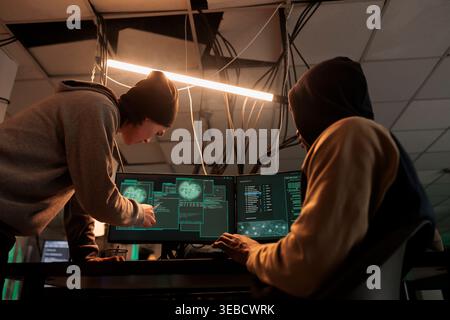 Hackers team hacking online server, stealing data, cybercrime. Cyberattack, it specialist pointing at code on pc screen, criminals programming internet virus at night time Stock Photo