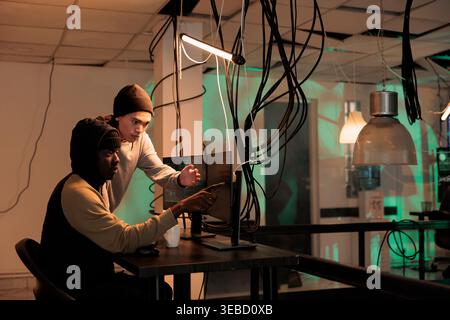 Diverse team of spies using trojan virus to ccause malware, working together to crack firewall encryption at night. Male hackers analyzing database to steal personal sensitive information. Stock Photo