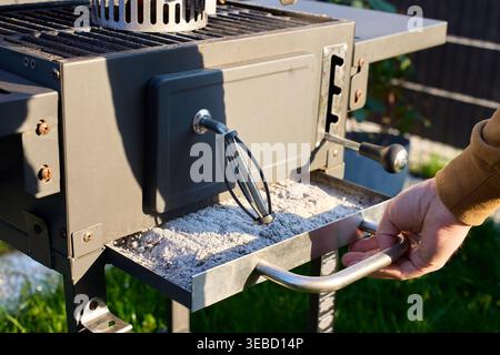 Hand pulls an ash tray filled with ash from outdoor grill. Concept of barbecue cleaning and grill maintenance Stock Photo