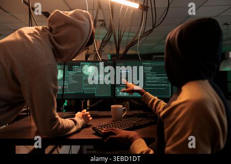 Internet criminals hacking online server, stealing finances, cyberattack. Hacker in hoodie pointing at code on pc screen, scammers programming malicious software at night time Stock Photo