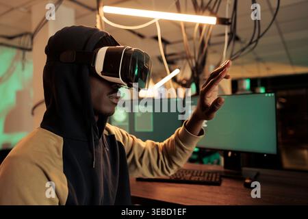 African american cybercriminal working with vr glasses at night, trying to break into government network with virtual reality. Male thief using interactive gadget to steal official information. Stock Photo