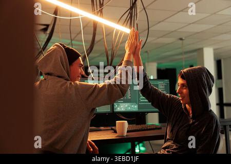 Successful cyberattack, happy hackers giving high five, hacking server, stealing business data. Scammers cracking computer system password, breaking into banking account at night time Stock Photo