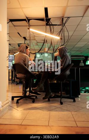 Young hackers coding trojan virus program to hack system, IT specialists programming illegal firewall encryption. Two cybercriminals developing malicious software late at night. Stock Photo