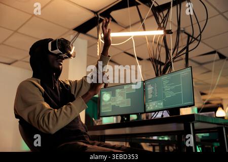 Hacker breaking into databse in vr glasses, spyware coding experience, african american man cracking computer system password in metaverse. Cybercrime, internet criminal developing malware Stock Photo