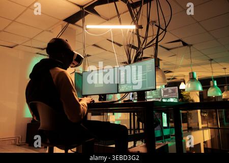 Male hacker working to hack server with vr 3d headset, planning cyberattack with virtual reality at night. Young adult causng computer malware, using interactive glasses, cyberterrorism. Stock Photo