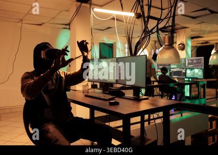 Male thief programming computer virus in vr headset, cracking database password in metaverse using interactive 3d glasses. Hacker coding malware in virtual reality gadget at night. Stock Photo